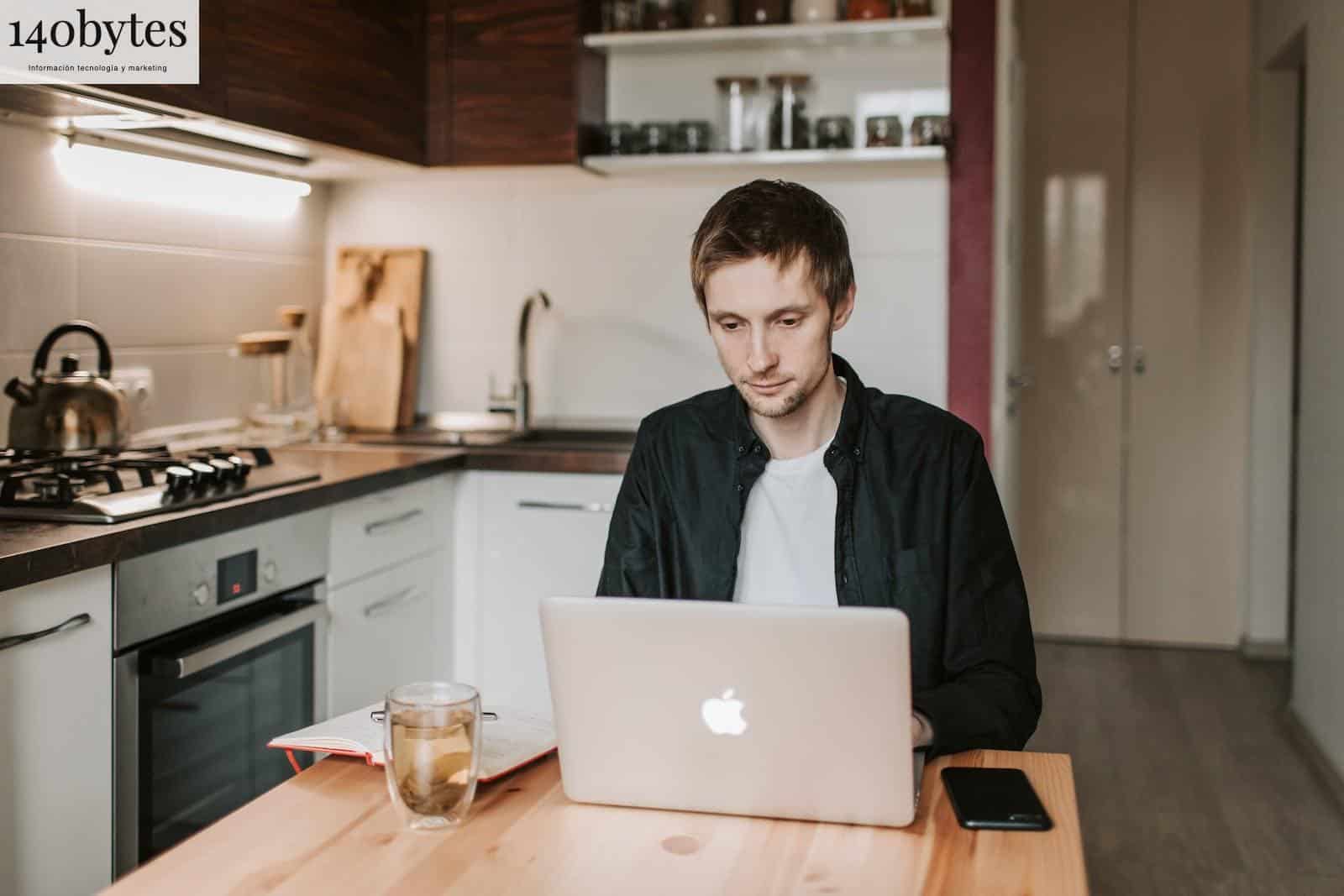 Pensive young male freelancer in casual clothes watching netbook while sitting at wooden table with glass of water and copybook in apartment